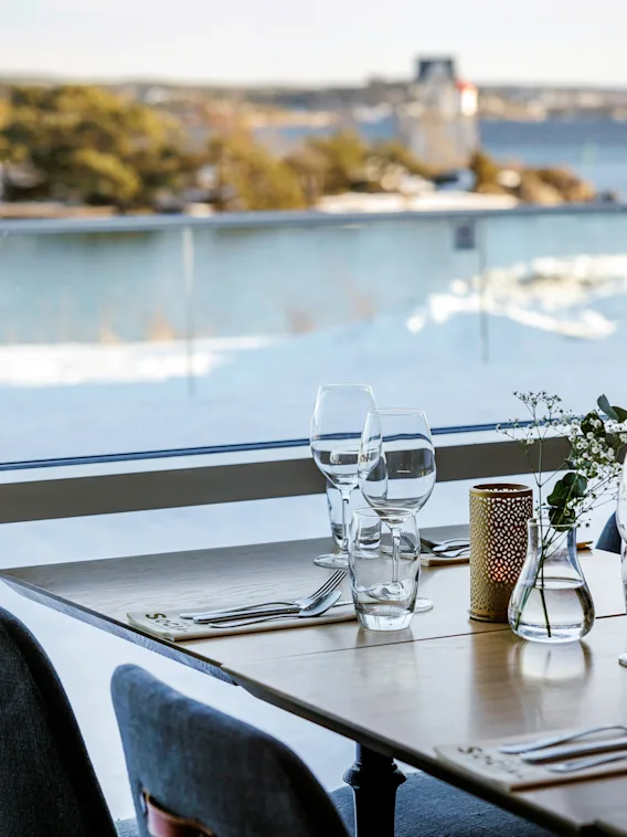 Ett träbord dukat för middag med glas och bestick har utsikt över ett snölandskap och en vattenyta genom ett stort fönster.