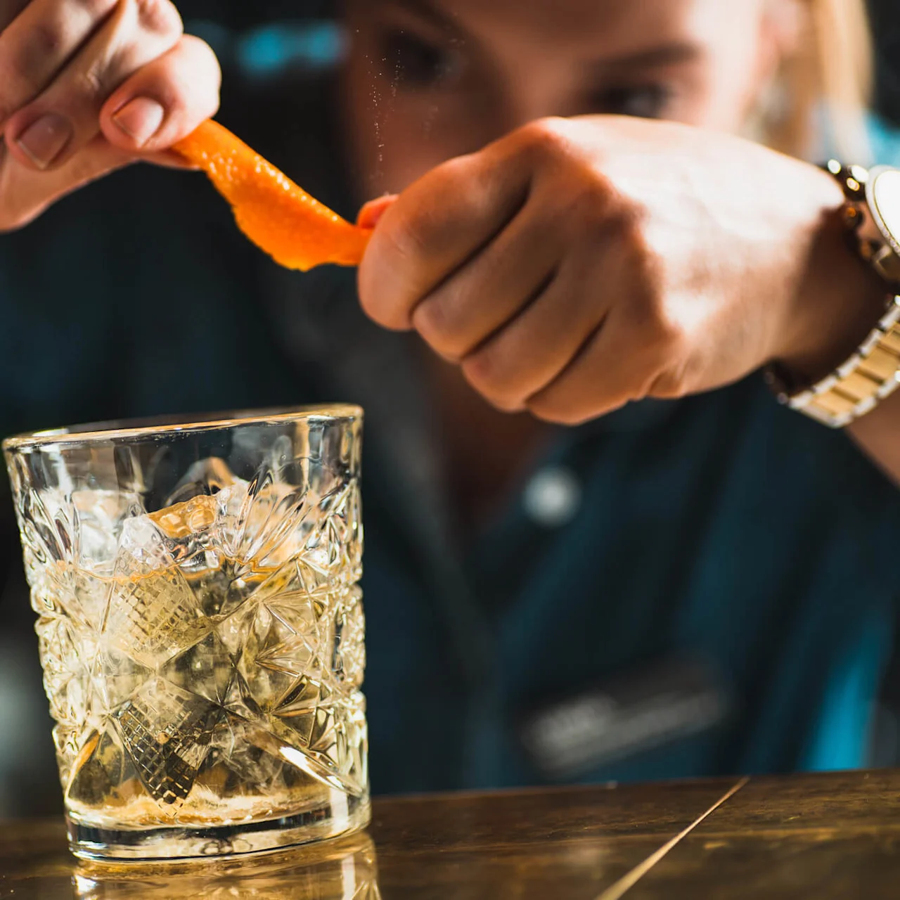 Et glas med en facetslebet overflade fyldt med is står på et bord. Hænder presser en appelsinskal over glasset, og olier frigives i drikken. Baggrunden er svagt oplyst.