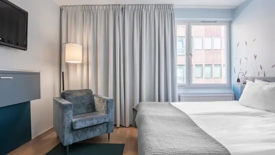 Ett mysigt hotellrum med en säng med vita sängkläder, en grå fåtölj, och en hängande lampa, beläget vid ett fönster med stängda gardiner. En väggmonterad TV finns.