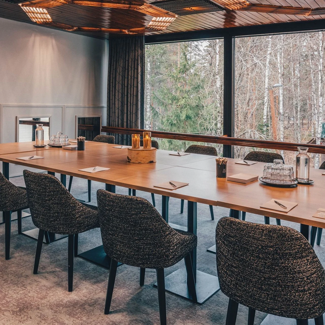 A long wooden conference table is arranged with chairs and notepads, set in a well-lit meeting room with large windows overlooking a forested area.