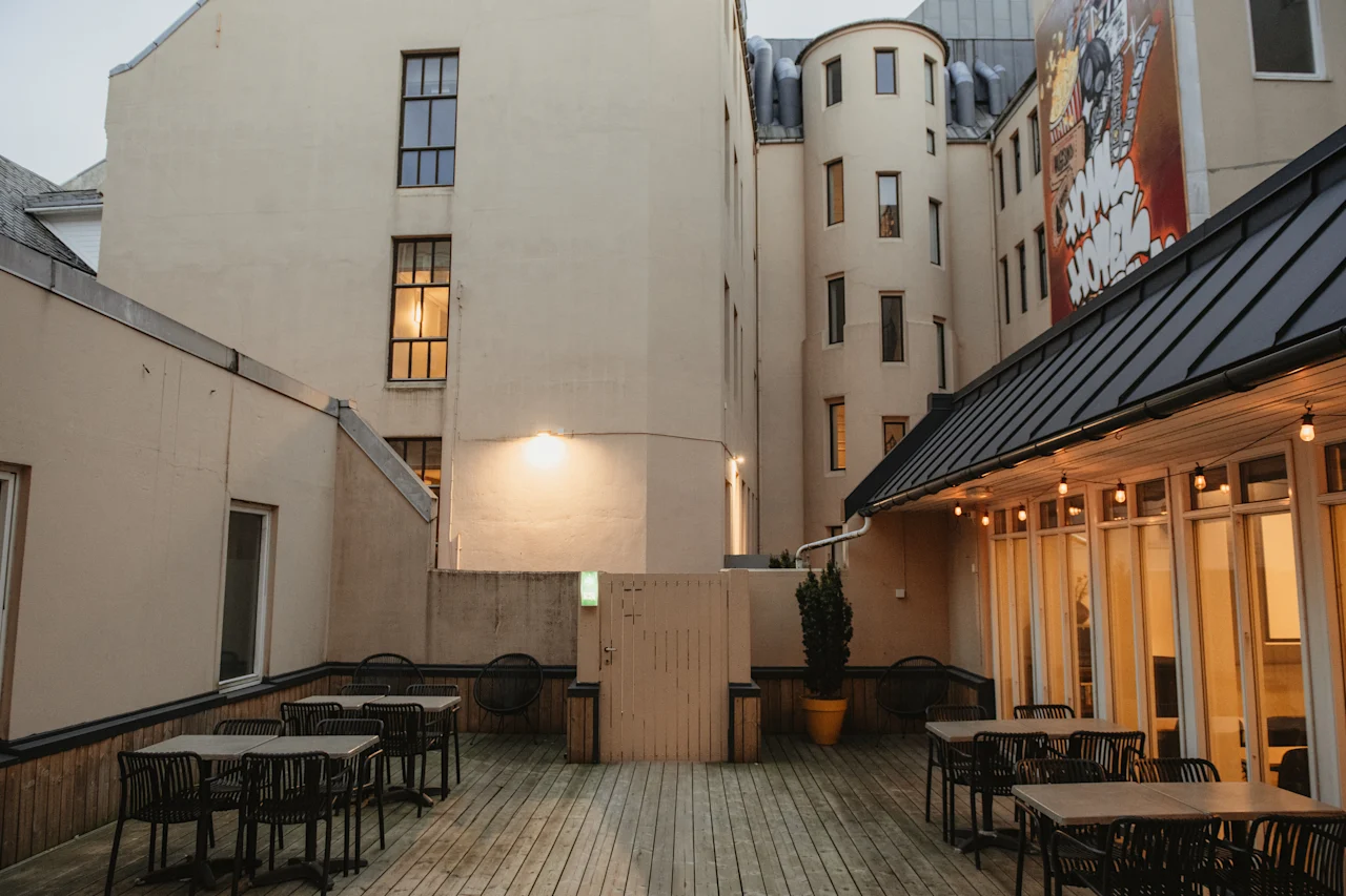 En indbydende udendørs terrasse med trægulv, borde og stole, omgivet af bygninger med varme lys og vægmaleri.