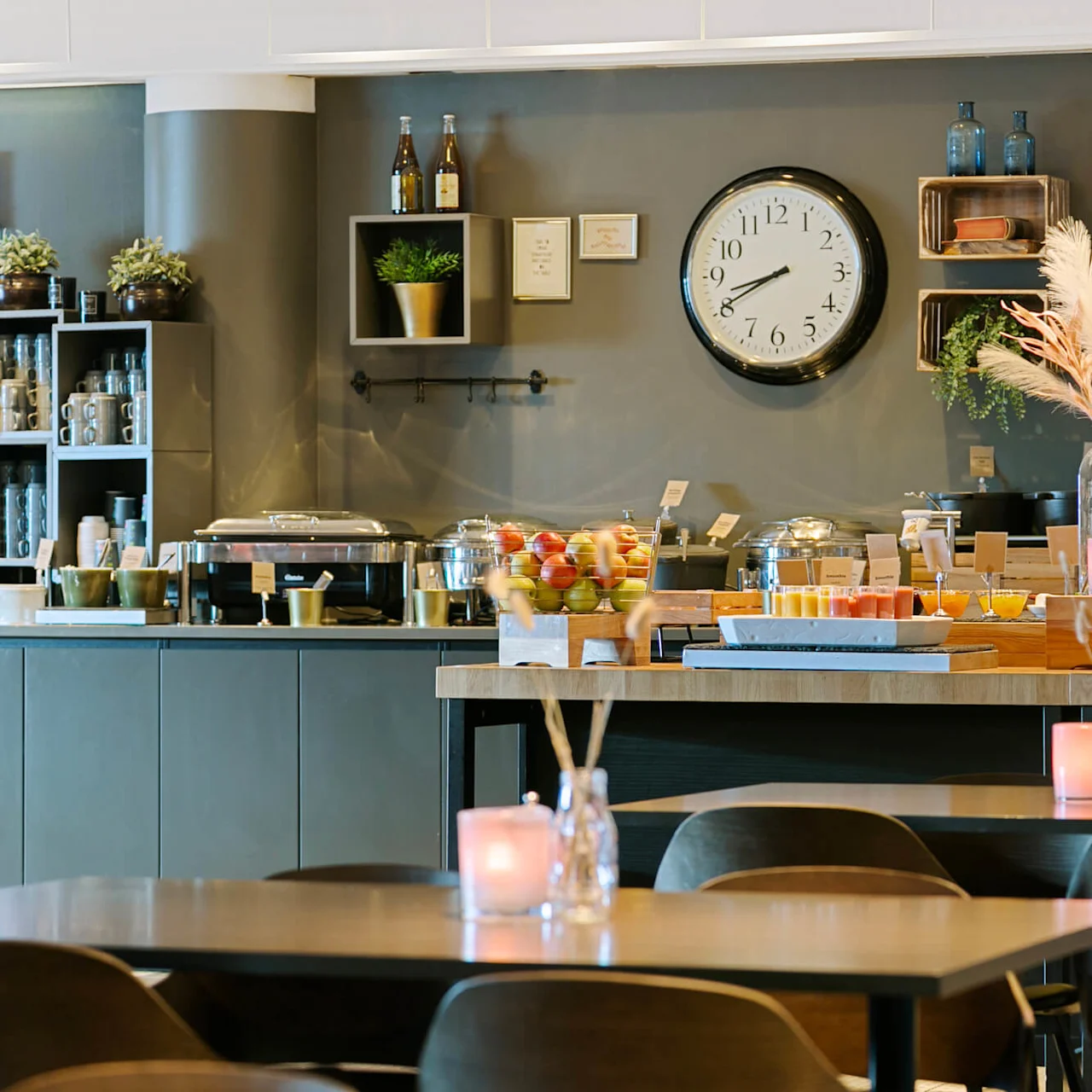 Cafeteria med en buffetopstilling med frugt, juice og salater på træborde. Bord med lys i forgrunden; vægur og dekoration i baggrunden.