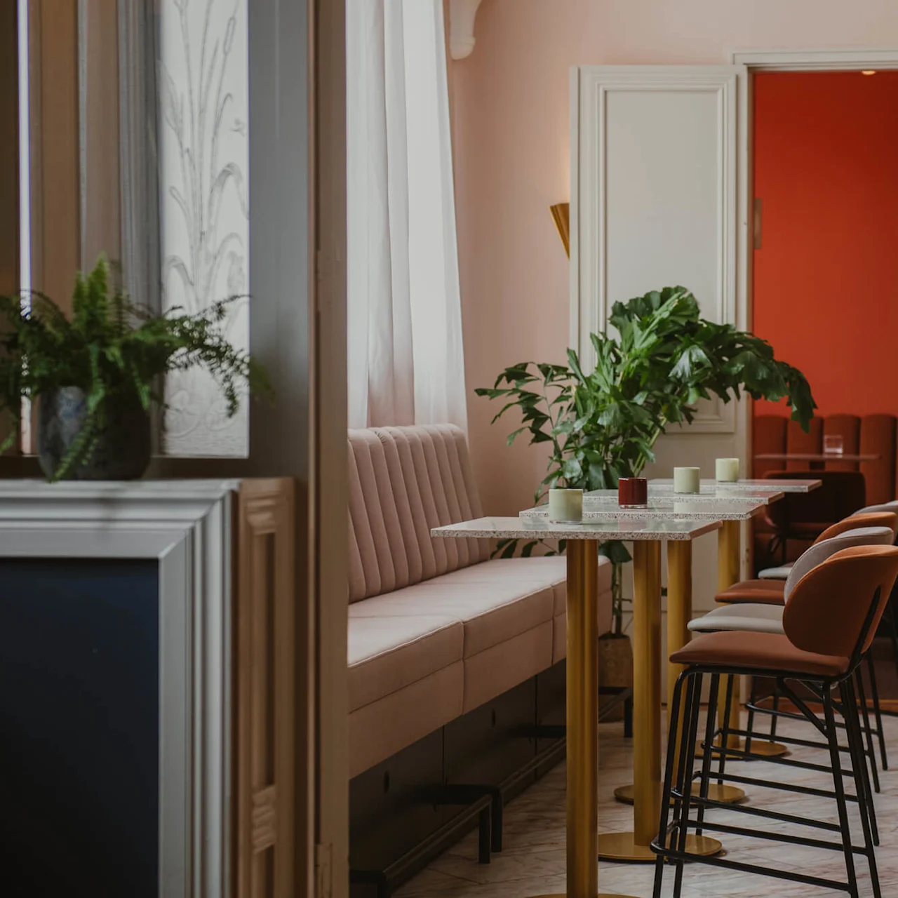 Tall tables with stools arranged alongside a pastel bench, plants decorating the space. A red-walled room is visible through a doorway, creating a cozy dining atmosphere.