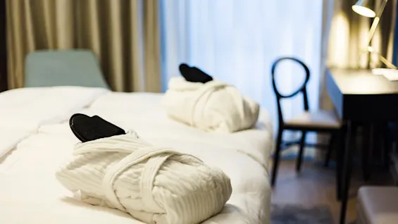 Bathrobes with slippers rest on a neatly made hotel bed; next to them, a chair and desk with a lamp create a minimalist, serene atmosphere.