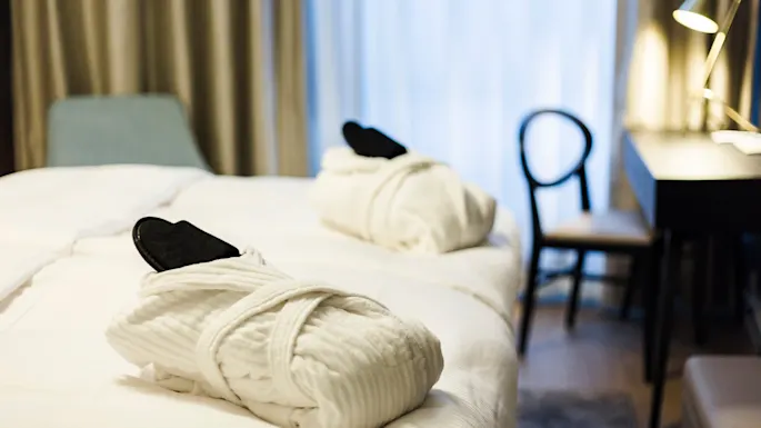 Kylpytakit ja tohvelit lepäävät siististi pedatulla hotellivuoteella; niiden vieressä tuoli ja lamppu kirjoituspöydällä luovat minimalistisen rauhallisen tunnelman.
