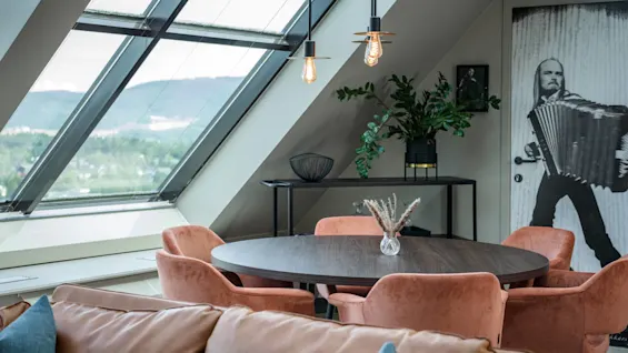 Et rundt trebord med en liten vase med tørkede blomster er omgitt av rosa stoler i en moderne stue med skråvinduer, en brun sofa og trekkspilltema på veggkunsten.