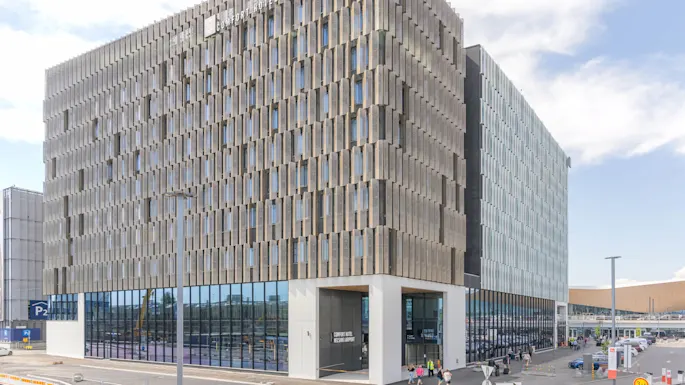 En moderne, geometrisk bygning merket "COMFORT HOTEL HELSINKI AIRPORT" står ved et parkeringsområde under en delvis skyet himmel. Folk går i nærheten.