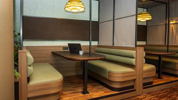 Cozy booth seating with a wooden table hosts a laptop and a mug in a warmly lit workspace. Modern pendant lighting and plush benches enhance the intimate office setting.