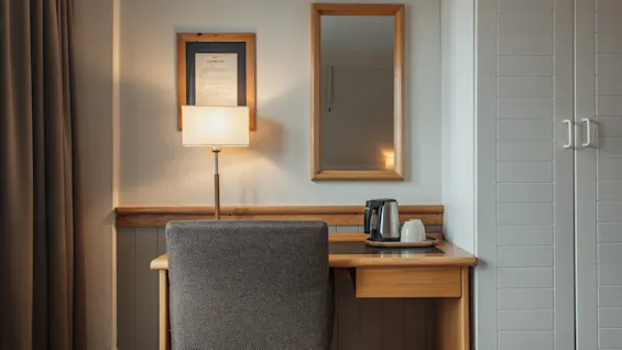 A cozy hotel room desk area with a lamp, mirror, and coffee making facilities, next to a wardrobe.