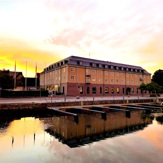En hotellbyggnad vid en hamn som speglar sig i vattnet under en gyllene solnedgång.