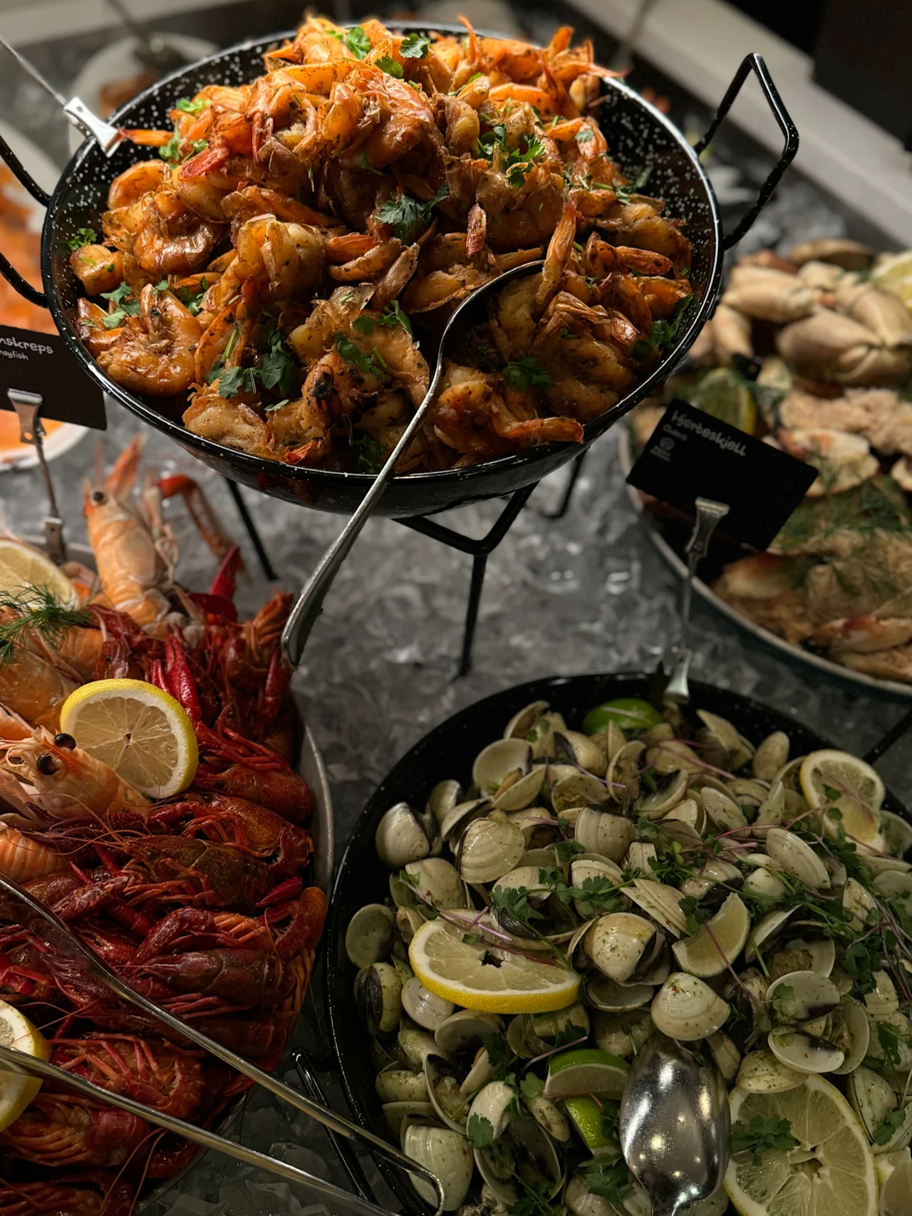 Fritert reker i en svart panne, garnert med urter, omgitt av kreps og muslinger på is. Små skilt med uleselig tekst følger med.