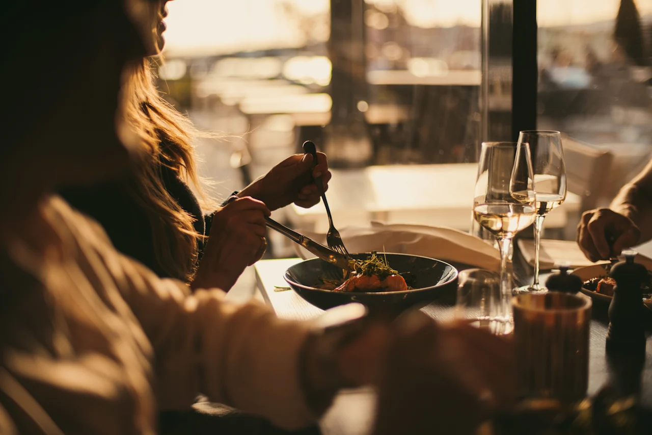 En person nyder et måltid med vin på en restaurant, med varm belysning og en sløret udendørsudsigt i baggrunden.