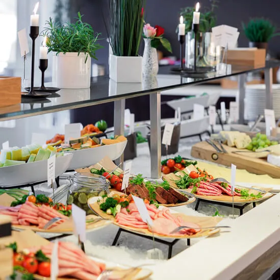 A buffet table displays an assortment of cold cuts, cheese, and fruits with decorative greenery and candles in a well-lit room. Wooden crate labeled "FARMERS MARKET" holds vegetables.