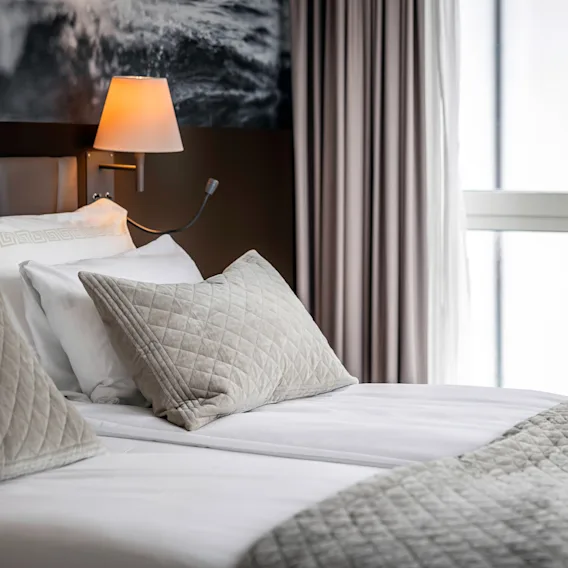En pent redd seng med quiltede puter og et grått teppe, plassert i et moderne soverom med en vegglampe og grå gardiner. Et svart-hvitt foto er på veggen.