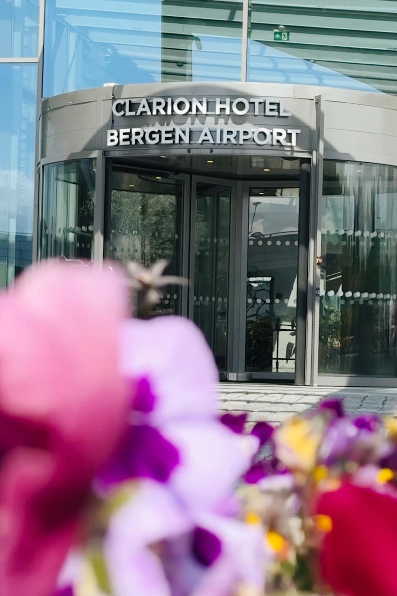 En roterende glassdør merket "Clarion Hotel Bergen Airport" med levende uskarpe blomster i forgrunnen, satt mot en moderne bygning.