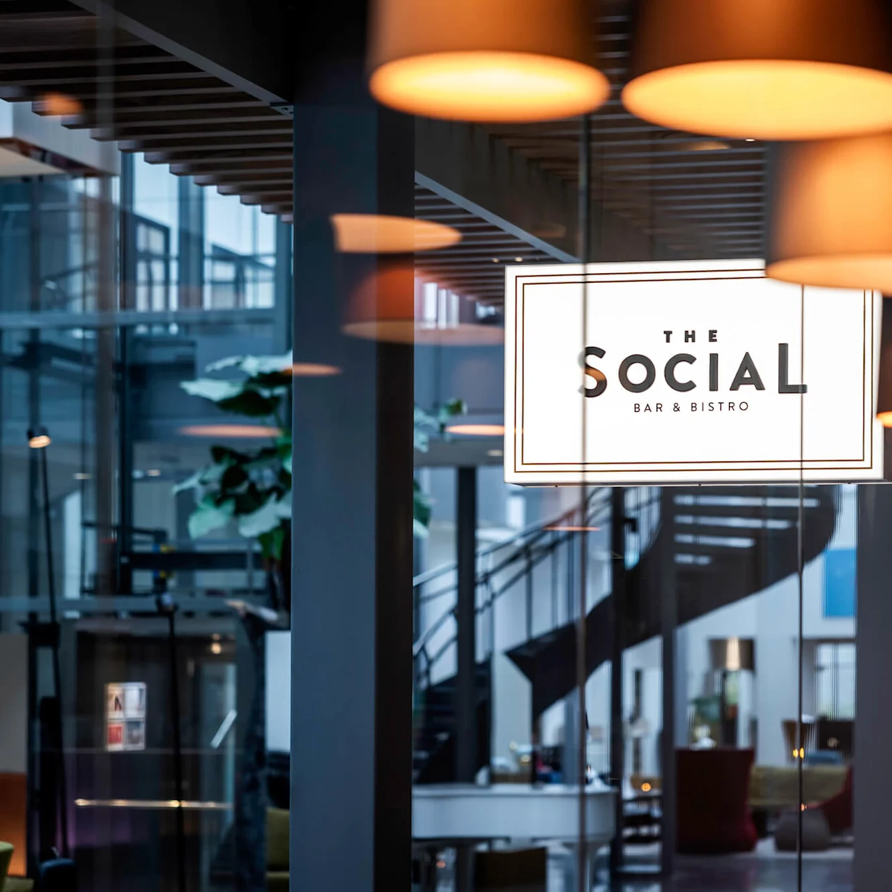 Lamper henger over et moderne innendørsområde med et skilt som lyder "The Social Bar & Bistro" på glass, som gjenspeiler den stilige atmosfæren. I bakgrunnen er det en trapp og planter.