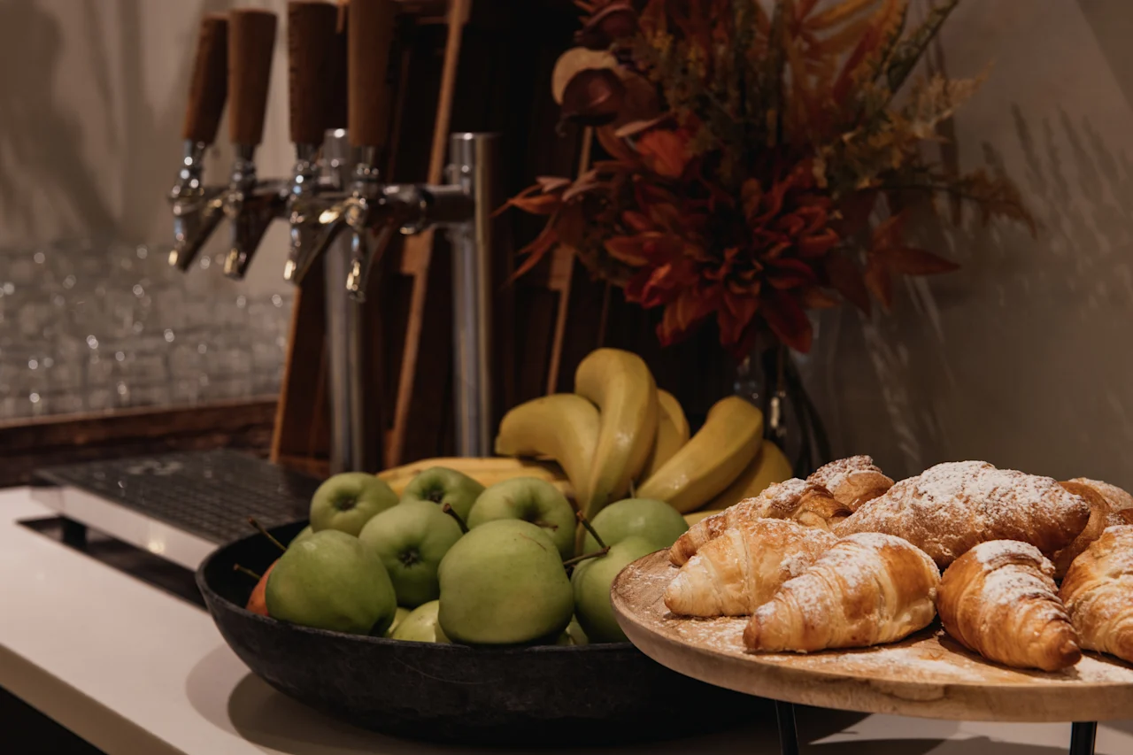 En morgenmadsbuffet med frisk frugt, croissanter og drikkedispensere, sat i en varm og indbydende ramme.