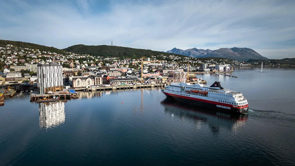 Ett stort passagerarfartyg seglar genom en hamn med en stad inbäddad mot kullar och berg under en blå himmel.