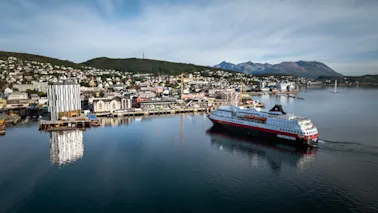 Et stort passasjerskip seiler gjennom en havn med en by som ligger inntil åser og fjell under en blå himmel.