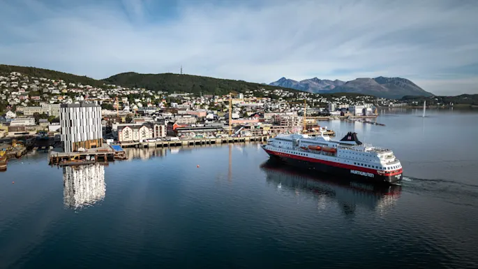 A large passenger ship sails through a harbor with a town nestled against hills and mountains under a blue sky.