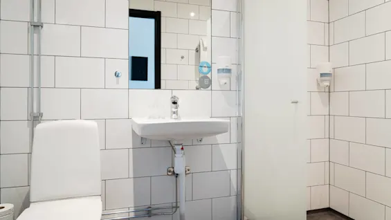En vit toalett och handfat i ett rent badrum, omgivna av vita kaklade väggar. En skylt lyder "DRINK MORE WATER". Dispenser och glas finns i närheten.