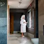 En person i badekåbe og håndklædehætte står ved et vindue i et spa, holder en kop, med en moderne pejs på en flisevæg i nærheden. Tekst: "minera velvære".