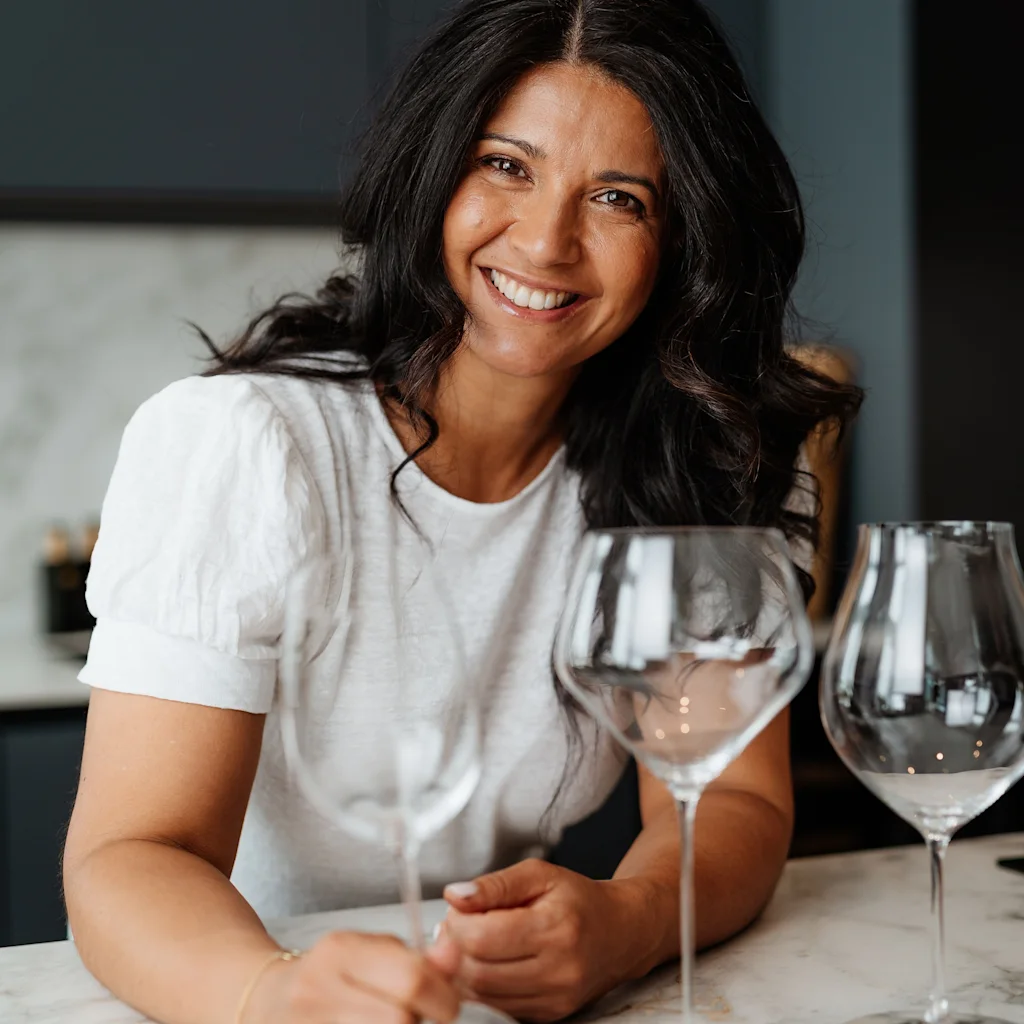 A smiling person holding a wine glass, with more glasses on a marble counter, ready for a social gathering.