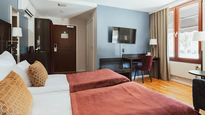 A tidy hotel room with two beds, a desk, and TV, illuminated by natural light from a window.