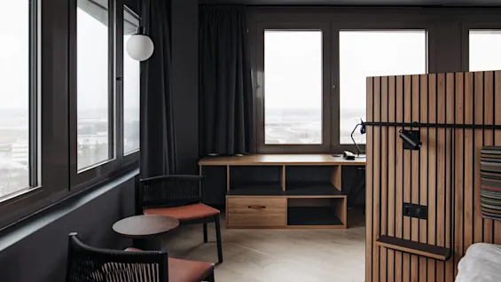 A wooden desk sits against large windows in a modern room. Two chairs and a small table are nearby, with dark curtains and a wood-panel partition adding to the sleek decor.