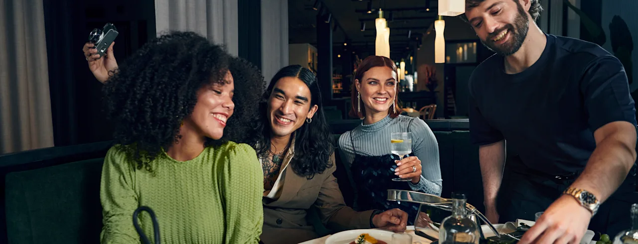 Clarion: Venner ler og deler et måltid på en livlig restaurant, med en servitør som presenterer deilige retter.