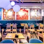 Restaurant interior featuring wooden tables and chairs, blue upholstered seating along the wall, and colorful artwork with abstract designs. Hanging globe lights add to the modern ambiance.