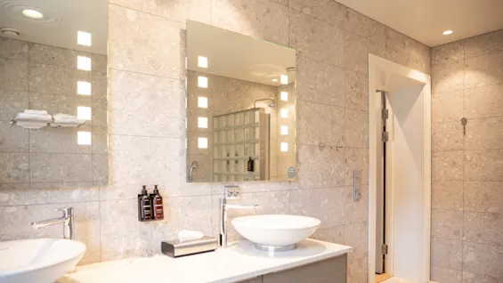 Two modern round sinks rest on a marble counter beneath illuminated mirrors. A tiled wall and doorway complete the bathroom setting, with toiletries neatly arranged between the sinks.