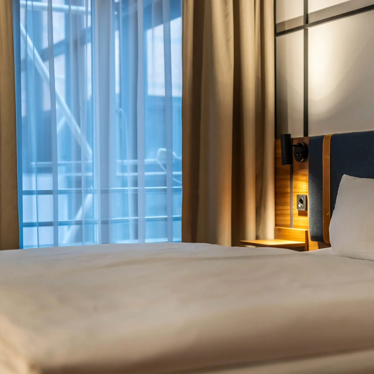  A close-up of a neatly made double bed with white bedding. The headboard is upholstered in blue fabric with wooden panel details. Behind the bed is a bedside table with a reading lamp, and large windows with beige curtains let in soft light.