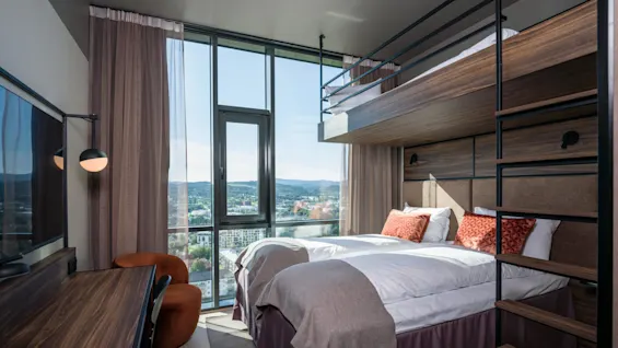 Et moderne hotellrom inneholder en dobbeltseng med oransje puter, en køyeseng ovenfor, et skrivebord med en stol, og store vinduer med utsikt over byen.