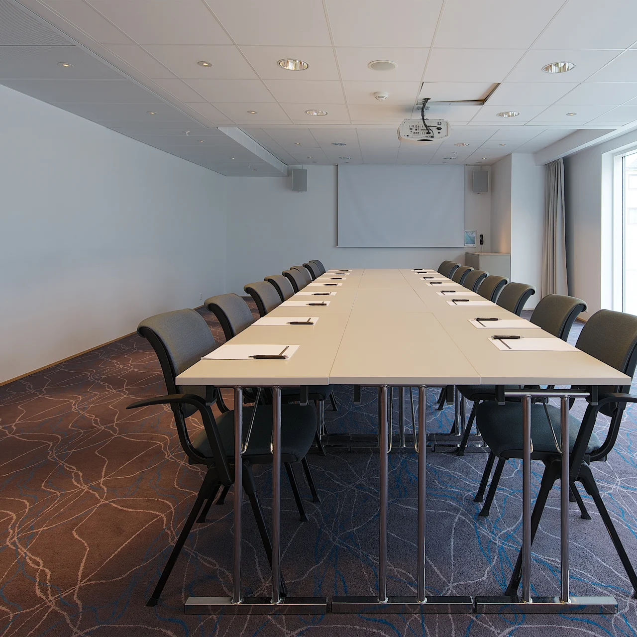 Långt konferensbord med anteckningsblock, omgivet av stolar och riktat mot en tom projektorduk. Ljust rum med mönstrad matta, vita väggar och stora fönster som släpper in naturligt ljus.