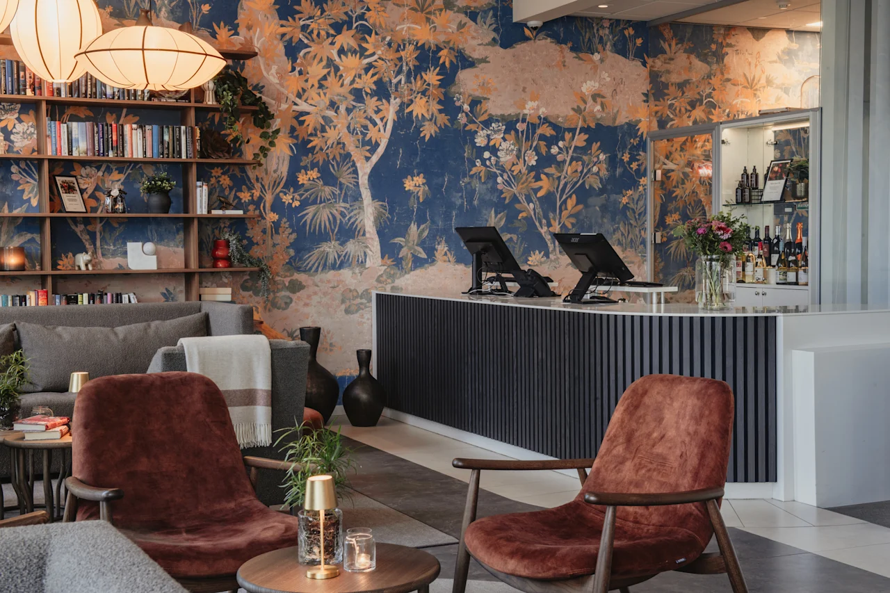 En hotellounge med servicedisk, bar og et hyggeligt siddeområde. Et levende vægmaleri af træer og løvværk pryder væggen.