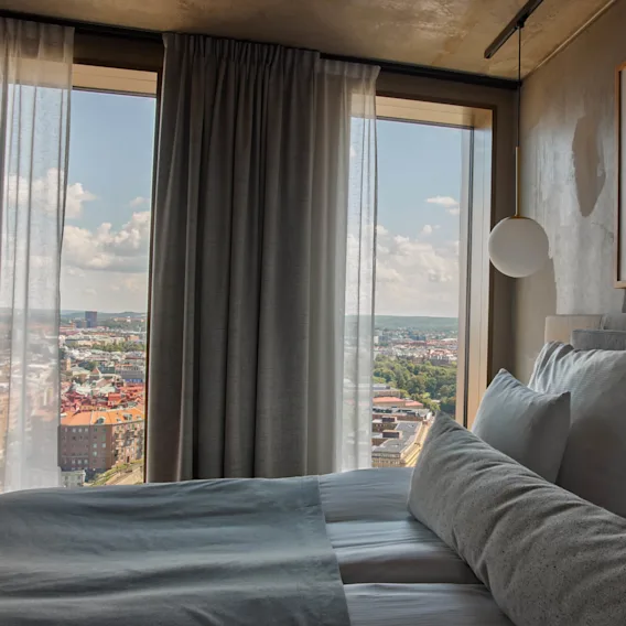 A neatly made bed with white pillows sits near large windows. A cityscape is visible through sheer curtains. Framed artwork hangs above, and a spherical lamp is suspended nearby.
