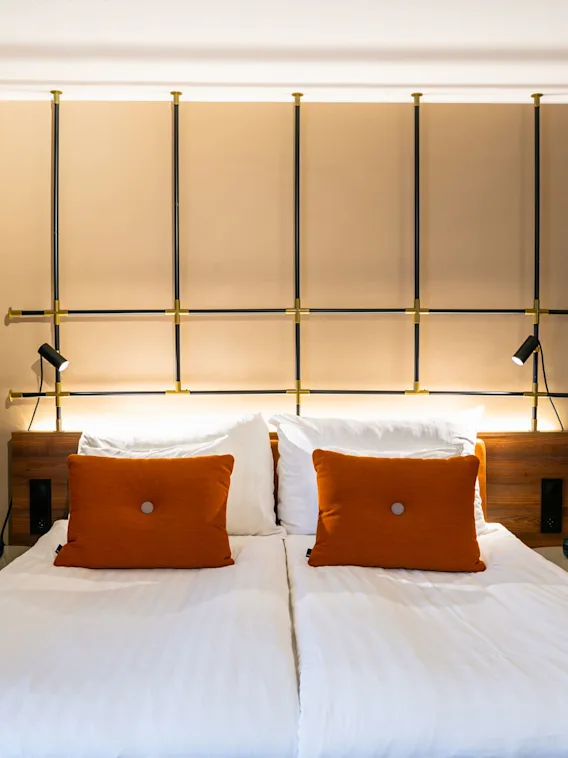 En prydligt bäddad säng med två orange kuddar står mot en vägg dekorerad med geometrisk design och små, riktade lampor i ett mjukt upplyst, modernt rum.