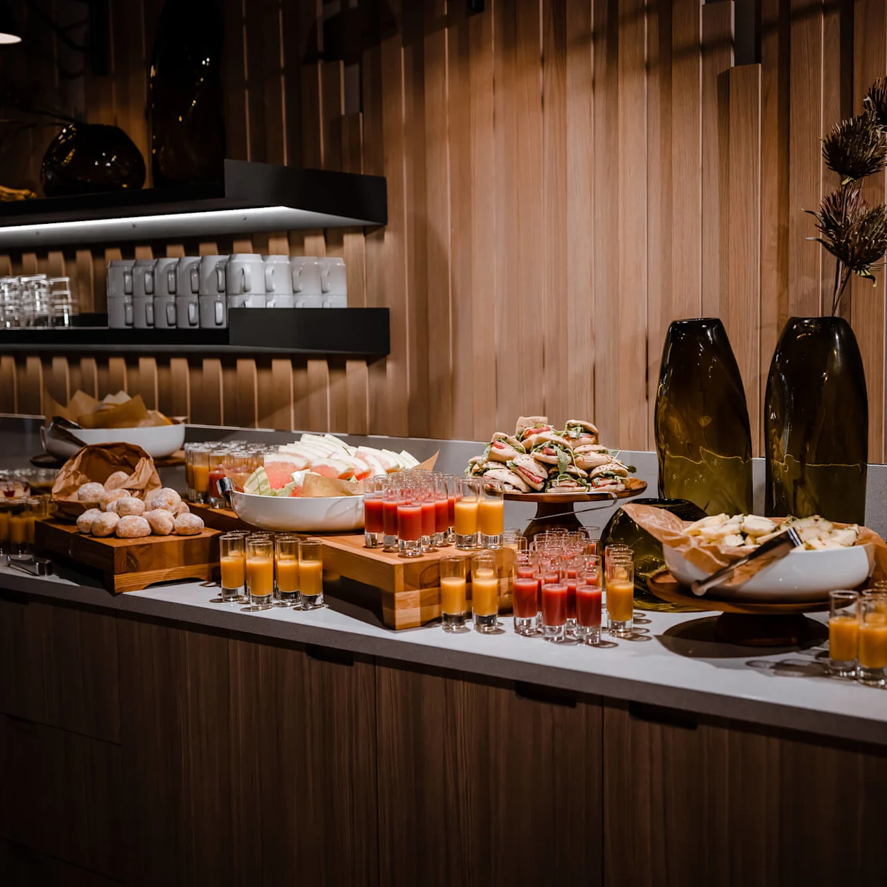 Buffet table showcasing various sandwiches, pastries, and fruit on wooden trays and plates. Small glasses of juice are arranged neatly. Wooden shelves with cups and glasses adorn the background.
