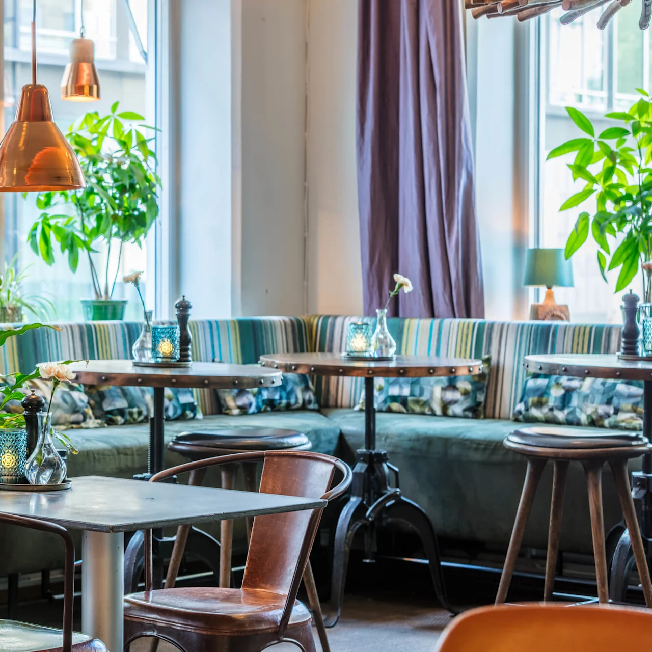 A cozy cafe interior with warm lighting, multiple tables, upholstered bench seating, hanging copper lamps, and potted plants giving a welcoming ambiance.