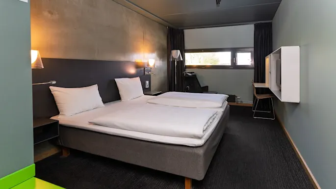 A neatly made double bed with white linens stands in a modern hotel room, featuring dark walls and a small desk near a window, illuminated by wall-mounted lamps.