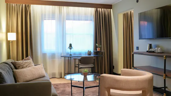 En moderne stue med en grå sofa og beige lænestol omkring et glas-sofabord. Et skrivebord med stol og lampe er ved vinduet, og et TV er vægmonteret.