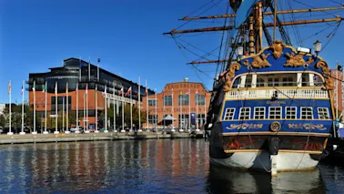 Ett stort blått och guld skepp vid namn Götheborg är förtöjt vid en kaj kantad av flaggor och moderna byggnader. I närheten syns "Eriksbergs Mek. Verkstads AB" på en tegelbyggnad.