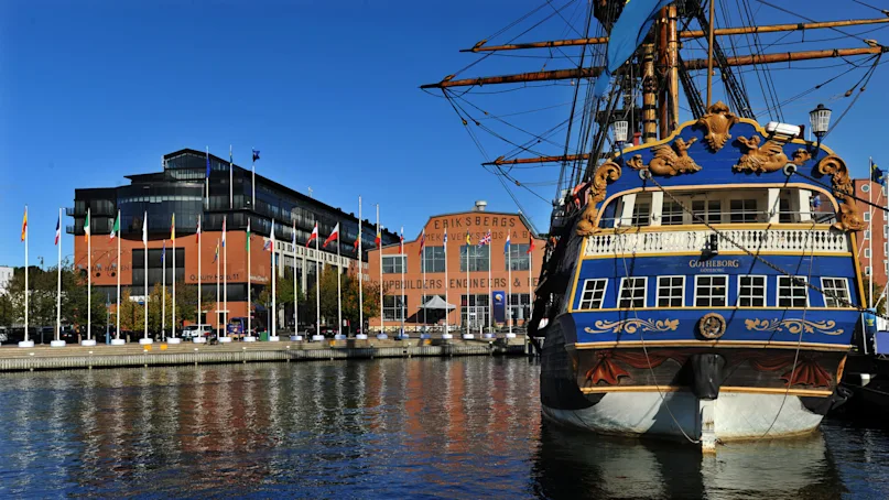 A large blue and gold ship named Götheborg docks beside a waterfront lined with flags and modern buildings. Nearby, "Eriksbergs Mek. Verkstads AB" is visible on a brick structure.