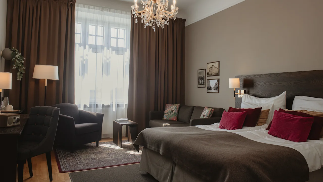 Ett mysigt hotellrum har en stor säng med vinröda kuddar, en ljuskrona ovanför och en sittgrupp med stolar vid ett gardinbeklätt fönster; varm belysning förstärker den inbjudande atmosfären.