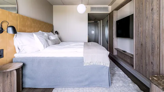 A king-sized bed with white pillows is neatly arranged. Two black wall lamps flank it. A round ceiling light hangs above, with a wooden wall and TV beside it in a modern hotel room.