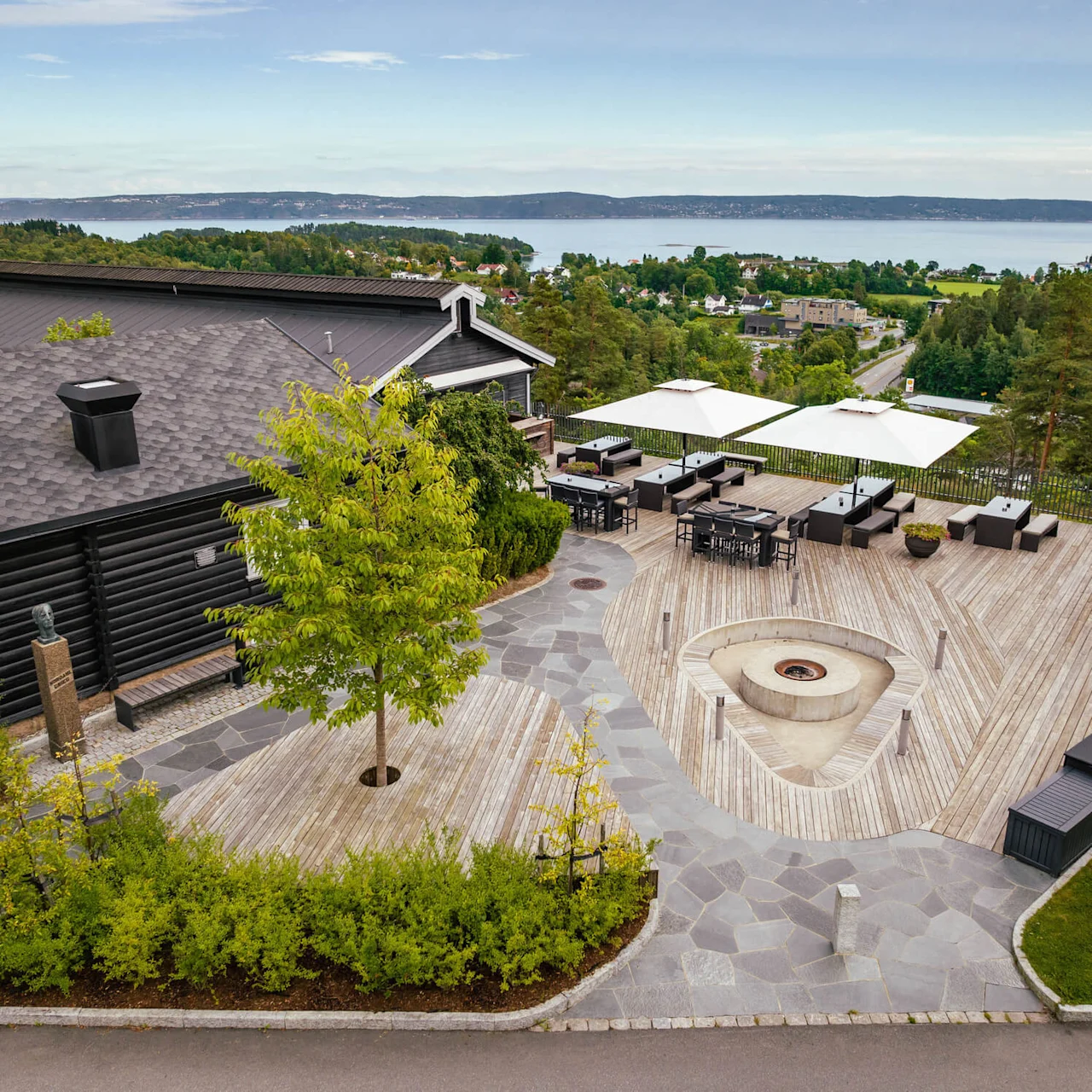 En romslig treterrasse med moderne utemøbler og en sentral ildsted. Omgitt av frodig grøntområde med utsikt over et naturskjønt landskap med fjerntliggende vann og bølgende åser.