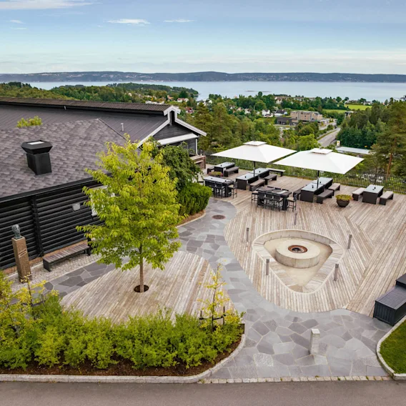 En romslig treterrasse med moderne utemøbler og en sentral ildsted. Omgitt av frodig grøntområde med utsikt over et naturskjønt landskap med fjerntliggende vann og bølgende åser.