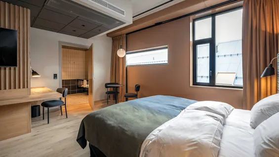 En seng står i et moderne hotelværelse, med et grønt tæppe og hvide puder. Ved siden af står et skrivebord i træ og en stol tæt ved badeværelsesdøren.