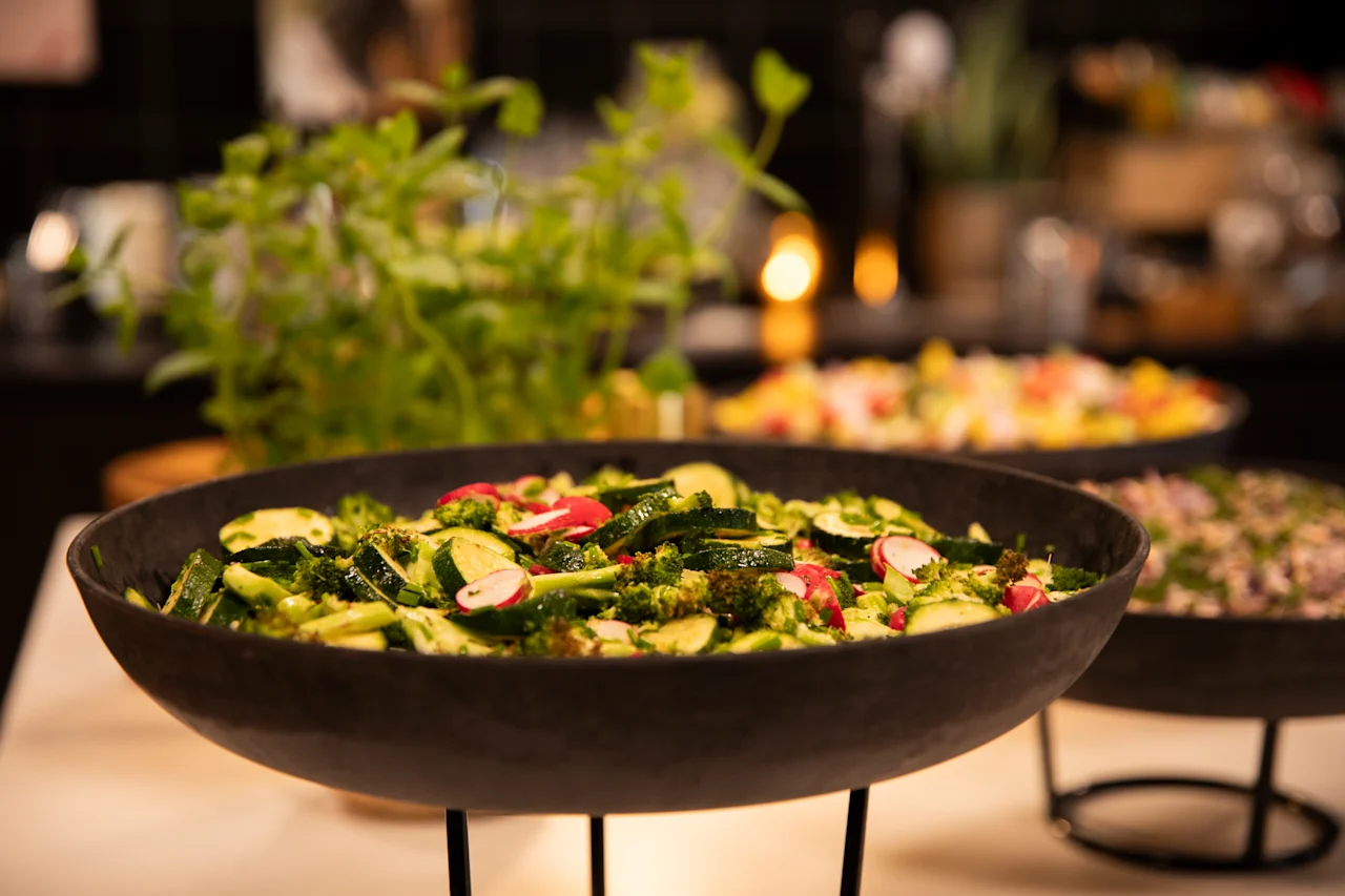 Et nærbilde av en frisk grønn salat med brokkoli, squash og reddiker i en mørk bolle på en buffet.